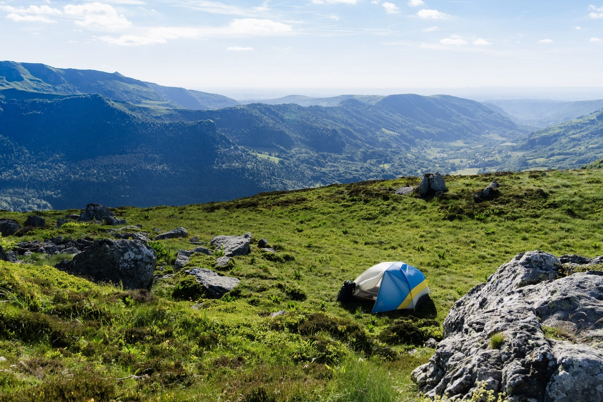 A small blue-and-yellow tent pitched among rocks on a grassy plateau, overlooking a wide valley in soft morning light