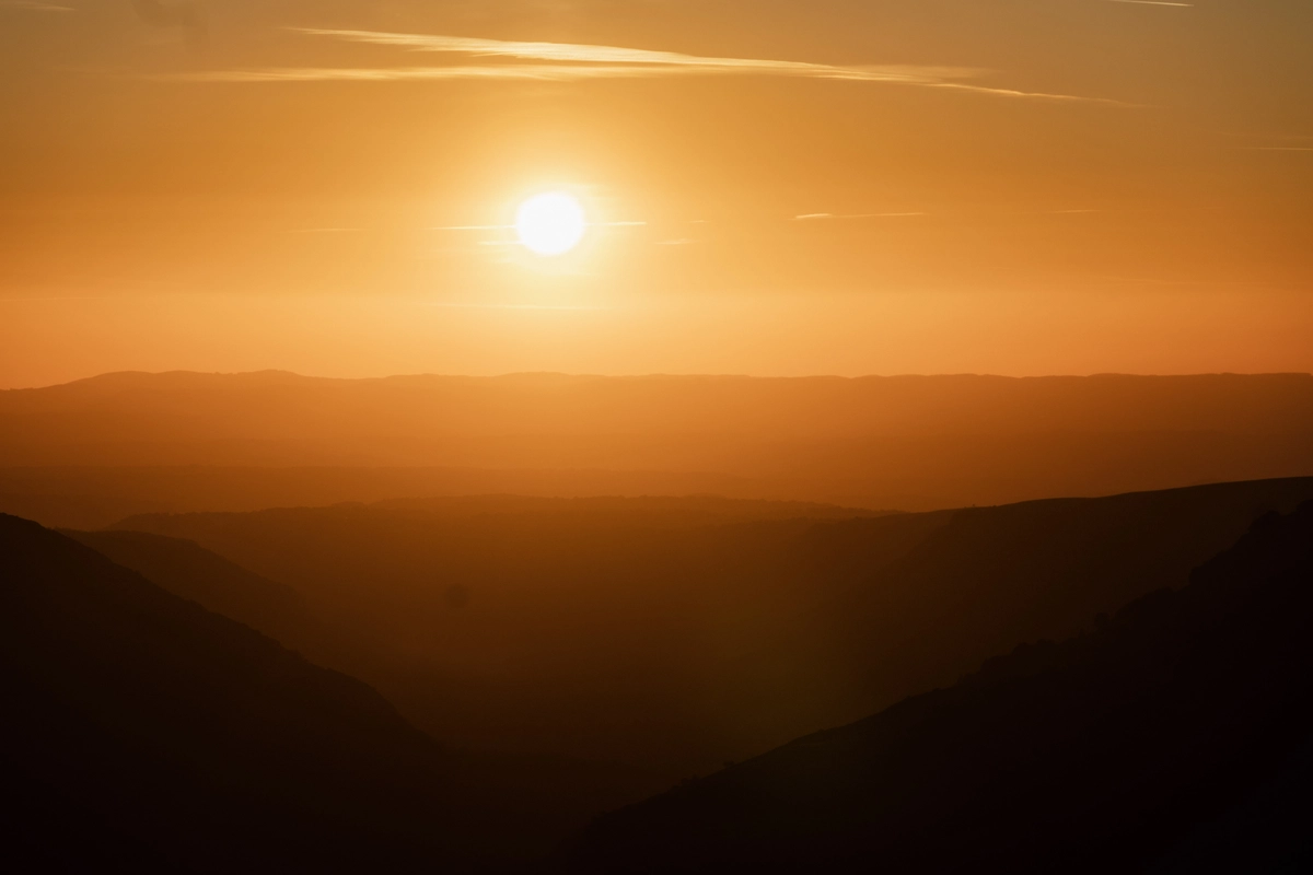 The sun setting into a warm orange sky over layers of hazy silhouetted hills and valleys