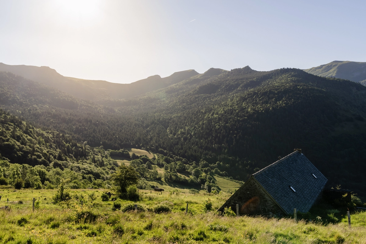 A slate-roofed stone farmhouse in a green forested valley, backlit by low morning sun over the surrounding mountains