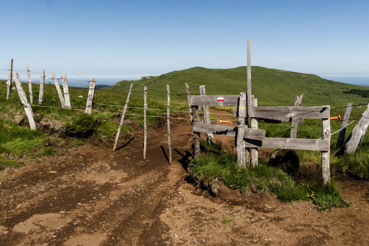 A wooden fence and stile on a worn dirt trail crossing open green highland pasture, marked with a red-and-white GR waymark