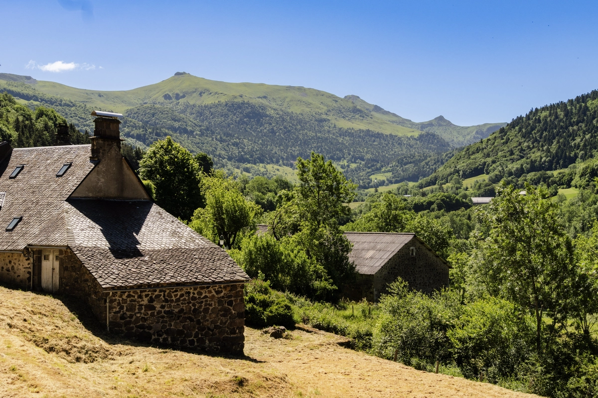 An old stone house with a tiled roof on a grassy slope, green wooded mountains and a valley stretching behind