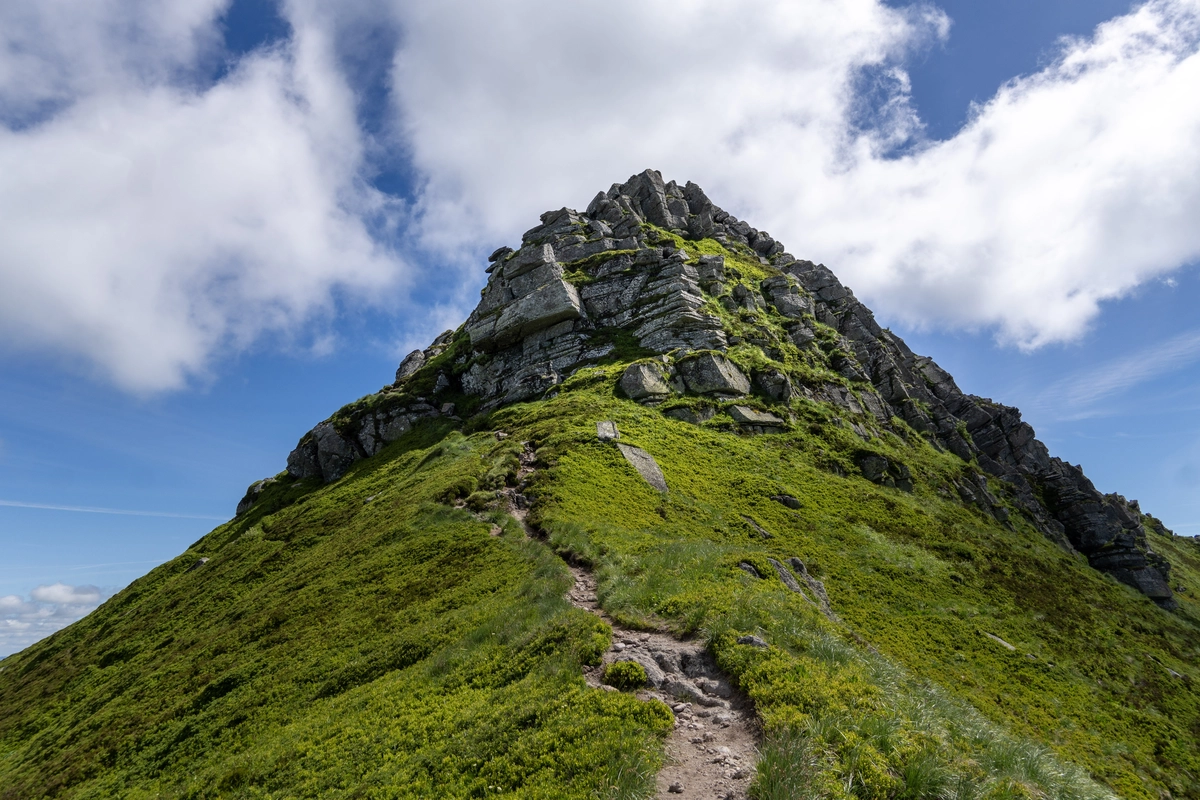 A jagged rocky pinnacle seen from below, a stony footpath climbing toward it under a blue sky with clouds
