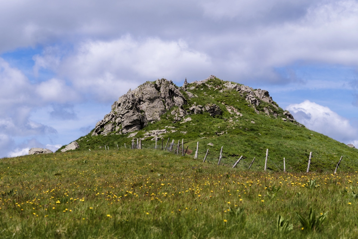 A rocky crag topped with a small cairn on a grassy hill dotted with yellow wildflowers, under a cloudy sky