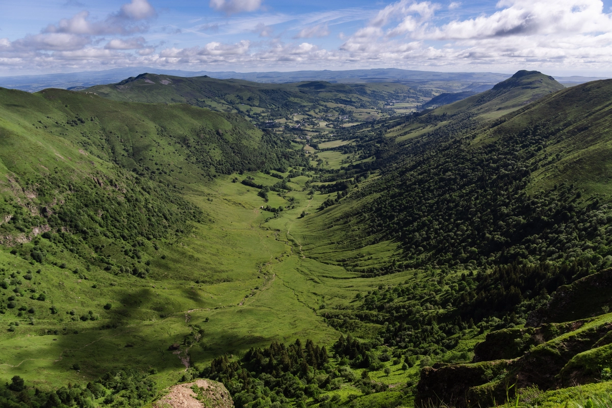 View down a deep, U-shaped glacial valley of green pastures from a high ridge, with a village and distant mountains beyond