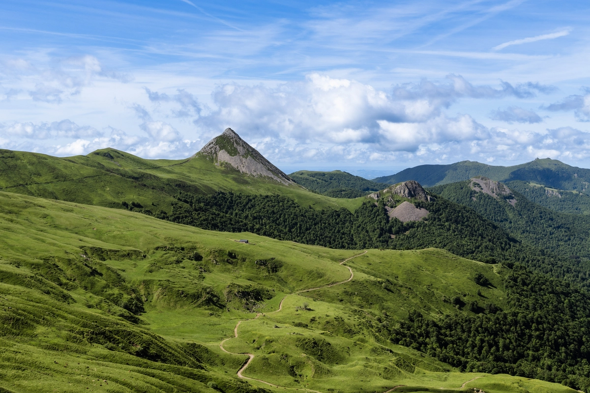 A sharp pyramid-shaped rocky peak rising above green volcanic slopes, a footpath snaking up toward it