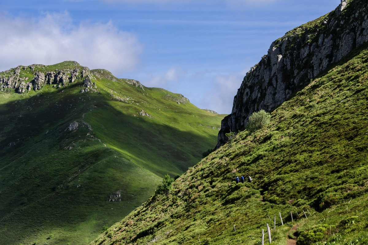 Two hikers in blue on a narrow path traversing a steep grassy mountainside beneath rocky cliffs