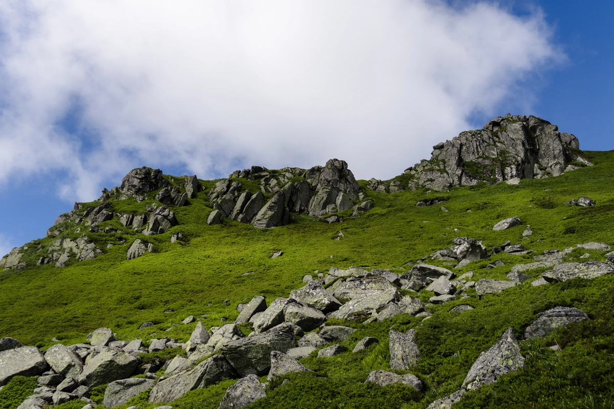 Weathered rocky outcrops and a boulder field scattered across a grassy summit slope under a bright cloudy sky