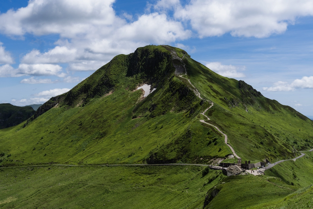 The broad green pyramidal summit of Puy Mary with footpaths and a small stone building at its base, under a blue sky with cumulus clouds