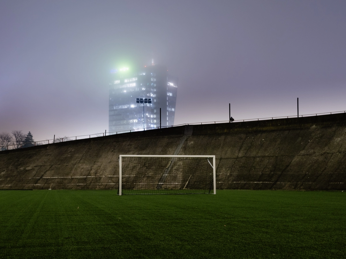 Empty football goal on a pitch with a glass office tower glowing in fog behind it